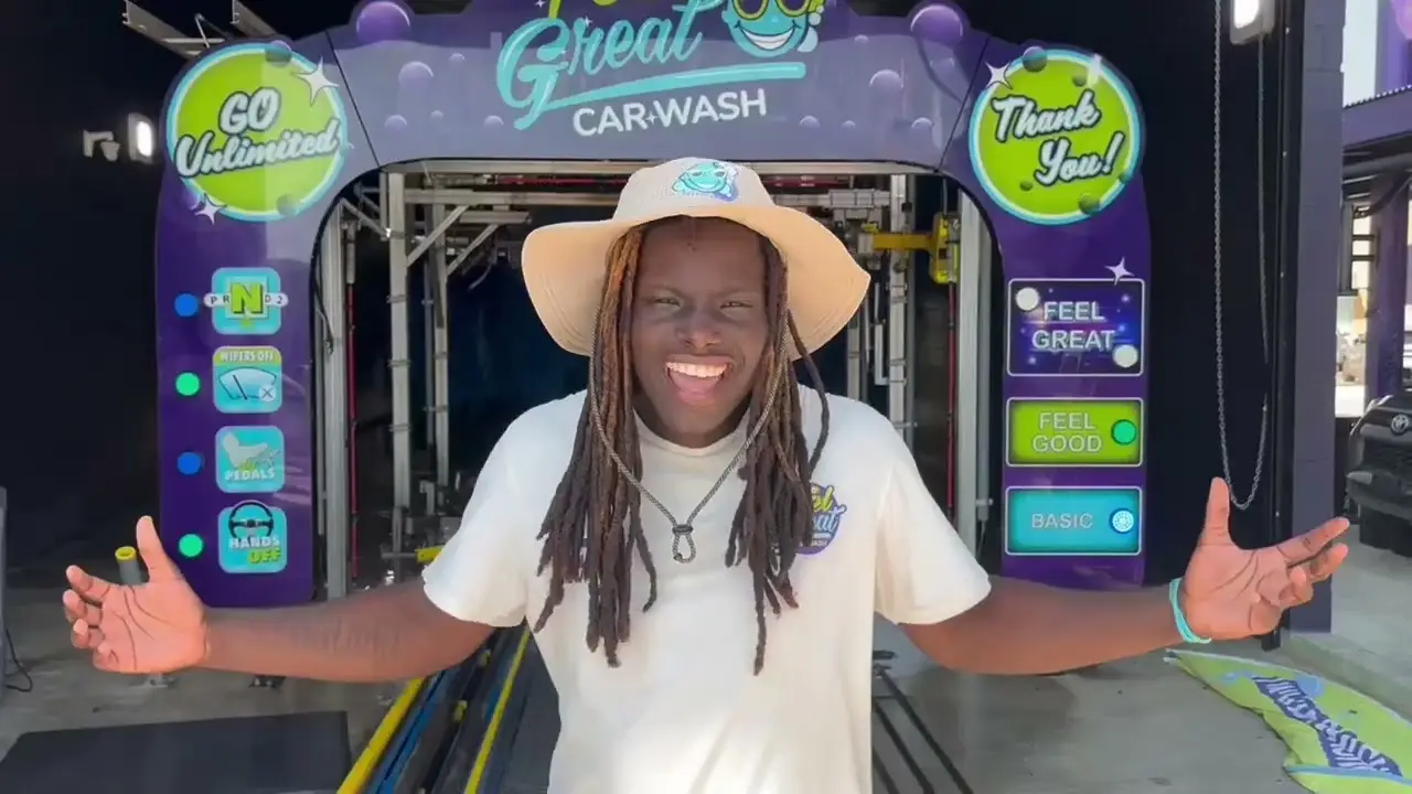 "A man with a large straw sun hat smiling and showing off the brightly lit 'Feel Great Car Wash' tunnel.